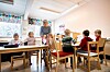 Barn och en vuxen vid bord i ett ljust förskoleklassrum med leksaker och papper.