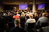 Publik i en konferenssal lyssnar till en presentation på scen med två stora bildskärmar.