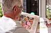 Äldre person läser en tidning med stora uppslag vid ett bord.