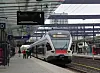 Ett tåg närmar sig perrongen på en täckt järnvägsstation med några väntande personer.
