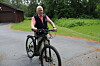 En man cyklar på en grusväg bredvid en träbyggnad och grön vegetation.
