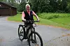 En man cyklar på en grusväg bredvid en träbyggnad och grön vegetation.