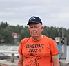 Man i orange T-shirt och mörk keps står vid en hamn med båtar och skog i bakgrunden.