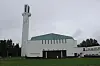 Modern vit kyrkobyggnad med högt torn, grönt tak och gräsmatta under mulen himmel.