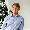 Porträtt av Ronnie Grandell i ljus skjorta framför en vit vägg och en grön växt.