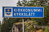 Blå vägskylt med texten Kirkkonummi Kyrkslätt och kommunens vapen framför skog.