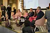 Paneldeltagare sitter på scen framför en projicerad presentation inför publik.
