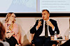 Paneldiskussion med en talande man i kostym, en lyssnande kvinna och en presentation i bakgrunden.