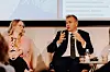 Paneldiskussion med en talande man i kostym, en lyssnande kvinna och en presentation i bakgrunden.