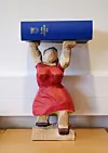 Träfigur i röd klänning som håller en blå skylt ovanför huvudet.