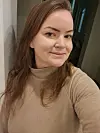 Kvinna med långt brunt hår i beige tröja poserar för ett självporträtt inomhus.