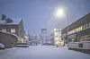 Snöig stadsgata med upplysta byggnader och gatlyktor under kvällens snöfall.