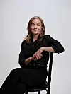 Leende kvinna i svart klädsel sitter på en stol mot en ljus bakgrund.